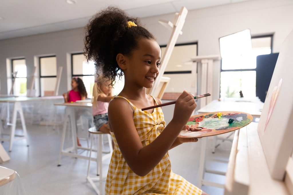 Smiling biracial elementary schoolgirl with palette and paintbrush painting on easel in school